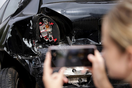 检查20多岁女驾驶员在事故发生后为移动电话保险索赔拍摄受损汽车照片的女司机后部图片