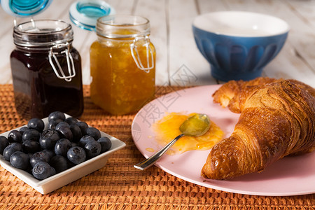 一种木制的早餐羊角面包蓝莓牛奶橙子和蓝莓果酱在桌布上方早餐牛角面包和蓝莓在桌布上方勺子图片