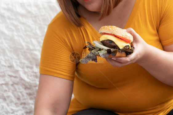 可口食物饥饿的超重妇女笑着拿汉堡包坐在卧室里她非常快乐享受餐吃狂食饮紊乱症概念芝士汉堡图片