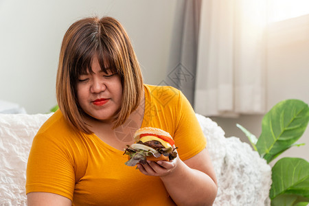 加大尺码快乐的饥饿超重妇女笑着拿汉堡包坐在卧室里她非常快乐享受餐吃狂食饮紊乱症概念一顿饭图片