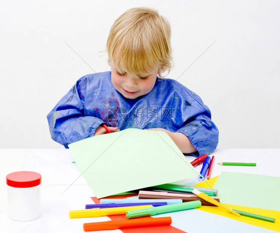 毛毡单身的年轻男孩快乐地切成一张纸板桌子上满了彩色铅笔胶水和有感觉的笔罐图片