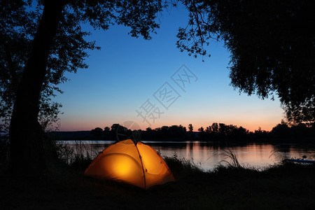营风景优美从湖岸的橙色帐篷内部照亮树木的轮廓日落天空夏风景自由和旅行的概念从湖岸橙色帐篷内部照亮树木的轮廓之中图片