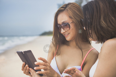 泰国读两个美丽的年轻女穿着比基尼在度假海滨旁的智能手机上寻找互联网图片