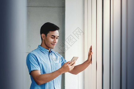启动联系男快乐的商人利用智能手机站在办公室窗口旁边通过移动电话和微笑阅读信息OfficeReadhessMessage图片
