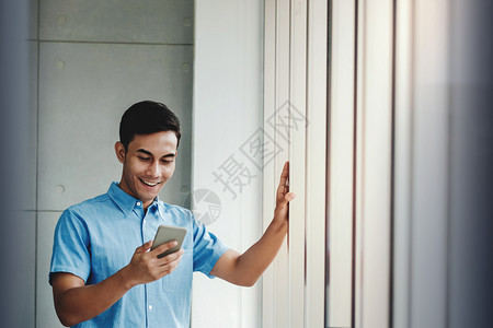 快乐的商人利用智能手机站在办公室窗口旁边通过移动电话和微笑阅读信息OfficeReadhessMessage窗户随意的技术图片