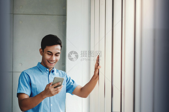 快乐的商人利用智能手机站在办公室窗口旁边通过移动电话和微笑阅读信息OfficeReadhessMessage窗户随意的技术图片