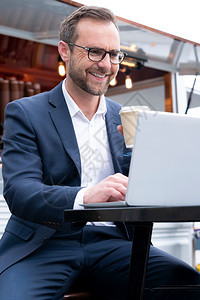通过户外咖啡店在笔记本电脑上工作的成熟商人喝着咖啡连接的垂直快乐图片