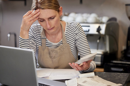 使用笔记本电脑和计算器通过账单寻找金融困难咖啡店的担心女所有者在财政困难中购买咖啡店票据铺水平的图片