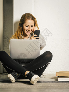 照片中一位年轻女坐在走廊上手持笔记本电脑笑着她的智能手机留言坐着在职的闲暇图片