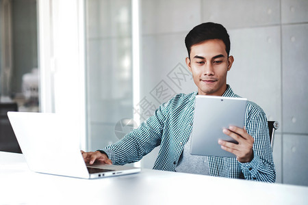 喜悦青年商人在办公室的电脑笔记本上工作微笑和看数字板现代的英俊图片