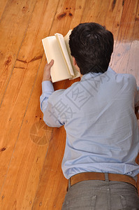 一名年轻人躺在家里地板上看一本商业书的背面景象学习木制的图片