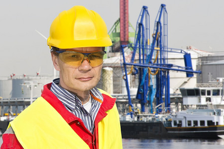 工程师筒仓阳光在一个石油化学港前在天然气设施无数起重机和一艘货船的石油化工港口前对一个自信的杜克工头进行肖像图片