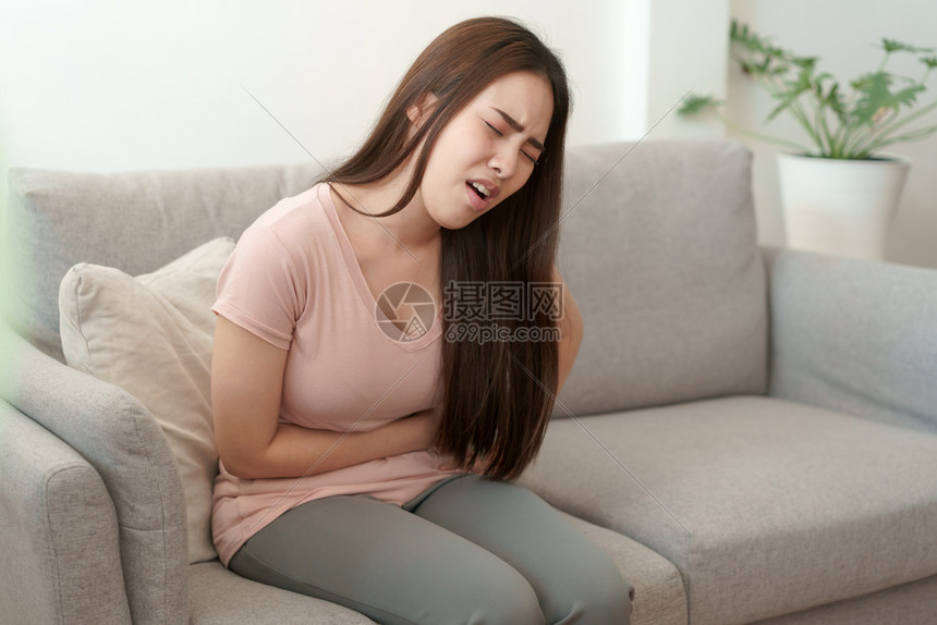 房间淑女遭受月经期和疼痛的亚洲女在家中坐沙发上痛苦地坐在沙发上令年轻女抽筋图片