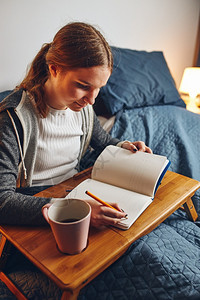 坐着做笔记阅读和学习的年轻妇女在隔离期间坐家中床上写作时从笔记本女孩写作杂志中读到年轻的文书工作图片