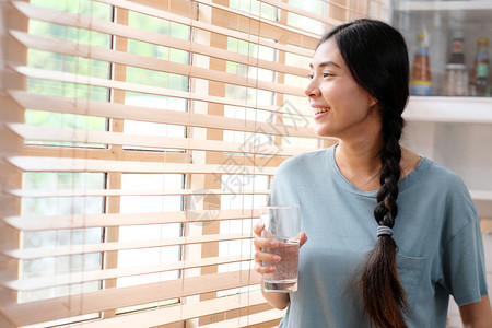 年轻美丽的亚洲女拿着一杯水笑微站在窗边坐厨房背景皮尔普和健康的生活方式窗边户饮食吸引人的图片