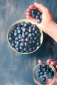 采摘浆果有机的女人把刚从罐子里收集到的蓝莓放一个小碗桌里在背景中漆成蓝色的桌子上图片