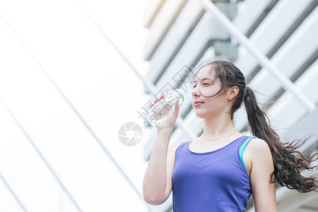 人们女士现代城市妇操控饮用水瓶运动时在现代城市中妇女穿着健康运动服在青年妇女户外健身锻炼以阳光明亮的为外面健康生活方式女概念现代图片