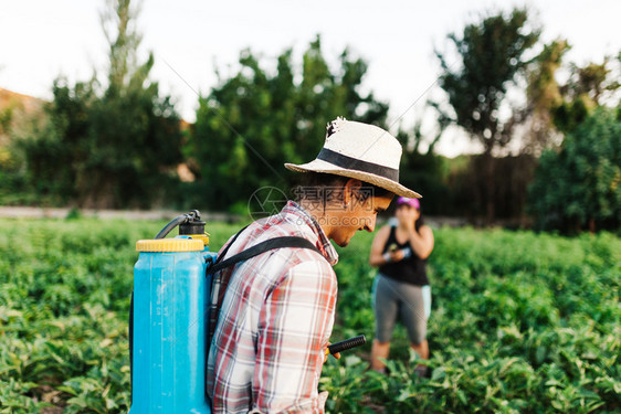 保护场地生物青年农民喷洒有机肥料身戴旧帽子和格衬衫的人工水泵罐图片