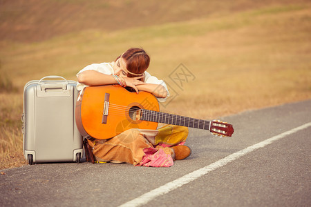 年轻嬉皮女青手持吉他和背包坐在农村公路上旅行漂亮的图片
