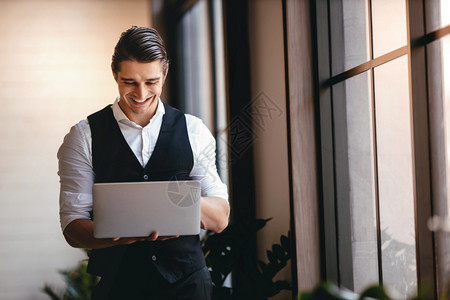 常设城市a从事现代工作场所计算机笔记本电脑工作的高加索青年商人在现代工作场所微笑的商人站在窗口前立男人图片