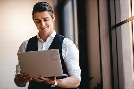 a从事现代工作场所计算机笔记本电脑工作的高加索青年商人在现代工作场所微笑的商人站在窗口前立信心雇员有创造力的图片