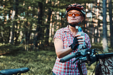 夏天活跃的女在森林里骑自行车度过免费暑假戴着自行车头盔和手套的女拿着带篮子的自行车从瓶子里喝水景观活动图片
