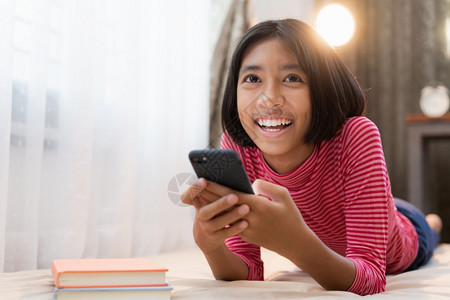 笔记本快乐的亚洲女孩在家晚上躺卧室里时使用智能手机亚洲孩子微笑并享受面部容表情概念放轻松家庭生活方式与自在学孩子们图片