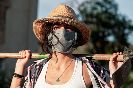 锄年轻农民妇女带着草帽走动的外科面具和蹄子望着照相机女户外图片