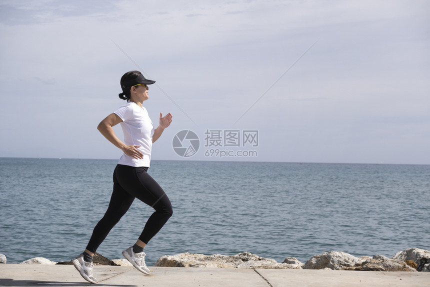 瘦身活力太阳镜戴帽子和太阳眼镜的女子沿着前台奔跑背景中的大海图片