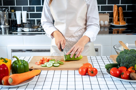 亚洲家庭主妇穿围裙用刀在切削纸板上黄瓜和番茄同时准备配料和烹饪供健康餐的午时在家厨房吃木板女士制的图片