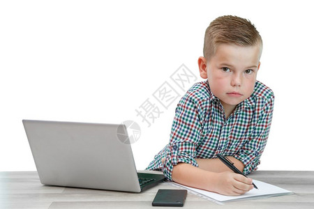 一种青少年身穿格子衬衫的高加索男孩坐在桌子上写一张纸上桌还有一台笔记本电脑和一部话那里图片
