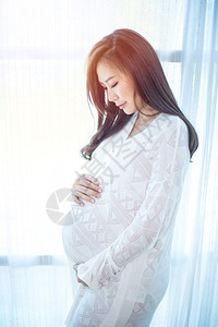 年轻的期待站在家窗户旁边的亚洲年轻美丽孕妇青年在家中的窗户旁边站着母图片
