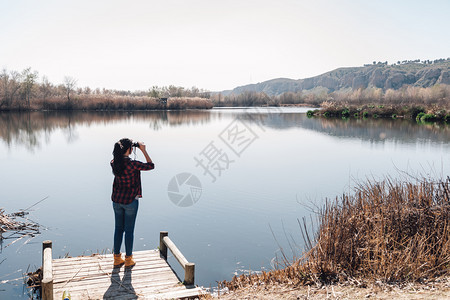 发现穿着双目望远镜和格子衬衣的湖角码头上黑发年轻女人旅游图片