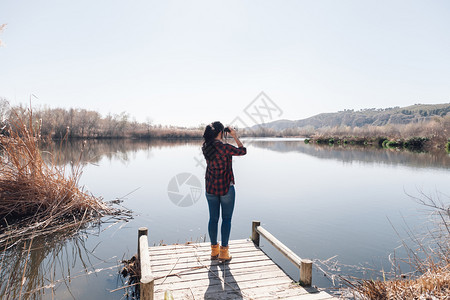 穿着双目望远镜和格子衬衣的湖角码头上黑发年轻女人背包客士探险家图片