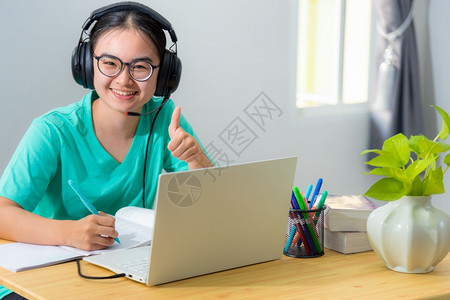 桌子有耳语眼镜的亚洲年轻女生肖像带着耳机眼镜的亚洲年轻女学生微笑着一个十几岁的女孩很高兴在大学网络远程研究上课中学习从家读笔记本图片