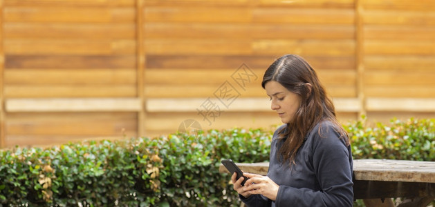 女士年轻的美丽西班牙裔年轻女子在早上坐公园长椅阅读手机的消息美丽西班牙裔年轻女子坐在公园长椅上阅读手机的消息快乐图片