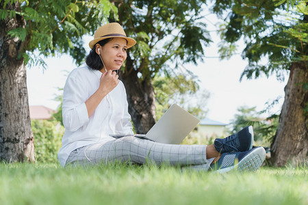 戴帽子的亚裔妇女坐在公园绿草地上使用带有线耳机的笔记本电脑肖像坐着尽管图片