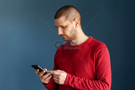 模型消息传递吸引人的白男子身穿红色毛衣站在蓝背景墙前使用智能手机发送短信或浏览互联网侧面视图的讯息Sms发短信或浏览视频图片