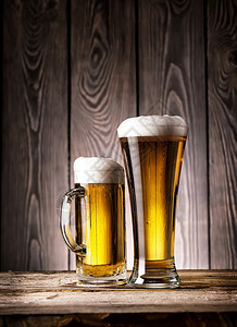 高压杯和一小轻啤酒木质背景上含泡沫的浅啤酒高压杯和一带有泡沫的轻啤酒马克杯小麦黄色的图片