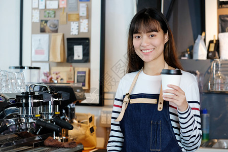 肖像在咖啡馆柜台背景小商业主食品和饮料业面笑脸的可支配咖啡杯中持有一次咖啡杯的年轻女咖啡会一种士图片