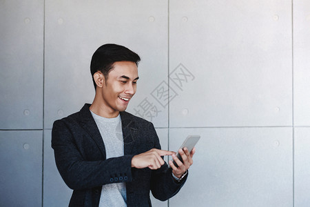年轻快乐商人的肖像使用智能手机站在工业混凝土墙边通过手机阅读信息和微笑年轻的幸福自信图片