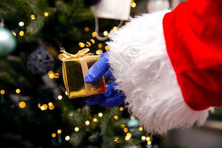 保持展示圣诞老人在树旁戴着手套和冠状面具拿着金色礼物Covid19和圣诞安全概念老人在树旁戴着手套和冠状面具拿着金色礼物Covi图片