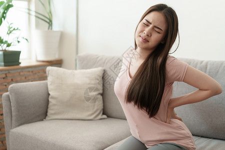 姿势年青女孩在家沙发上坐着时有瓦ist痛的亚裔女孩因肌肉健康和疾病概念而痛苦背部手图片