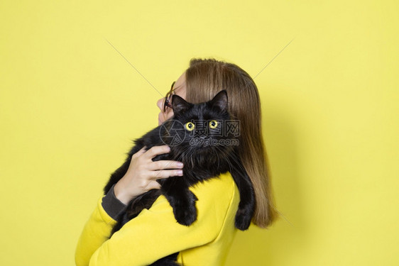 猫咪穿黄色连衣裙的女孩抱着她心爱的黄色背景上眼睛毛茸的黑猫宠物护理概念穿着黄色连衣裙的女孩抱着她心爱的黄色背景上眼睛毛茸的黑猫宠图片