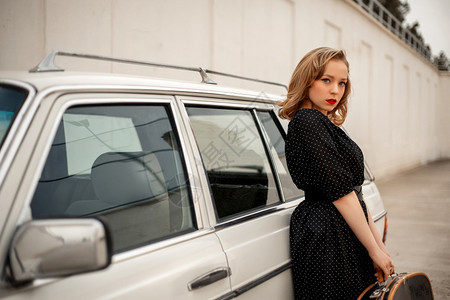 身着黑色古老的Polka点装饰的年轻美女坐在一辆旧白色汽车旁边手里拿着一个旧式手提箱车辆优质的年轻图片