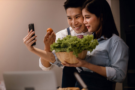 年轻的快乐移动a在烹饪自拍厨房或通过智能手机现场流动时使用电话的一对夫妇微笑同时准备在家中烹饪织女食品现代健康生活方式概念家庭图片
