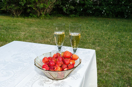 夏令周年纪念日碗加新鲜草莓和两杯玻璃在桌边上闪亮着葡萄酒花园里有白布饮料图片