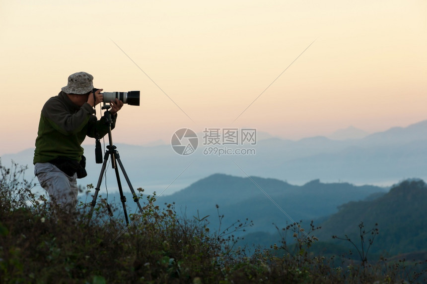 在日出时山峰的三脚架上用照相机和远光放大镜头拍摄照片的一位老年人侧面视图长焦思考游客图片