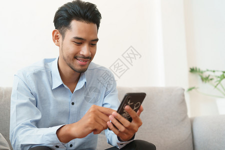 现代的穿着蓝衬衫年轻亚洲男子身着蓝衬衫其肖像正在用智能手机阅读坐在家里客厅沙发上吸引人的在线图片
