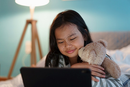 药片微笑女亚洲小孩在家中晚间卧室的数字平板电脑上躺着观看娱乐的亚洲女孩带着子快乐的笑容和拥抱图片